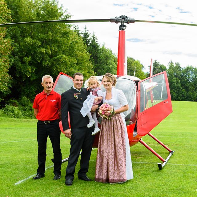 Ein Brautpaar mit einem Kind vor einem roten Helikopter auf einer Wiese