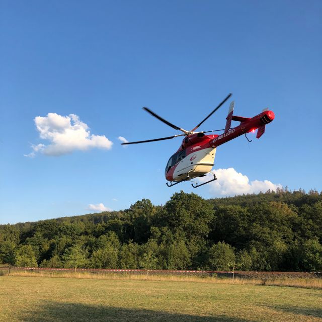 Ein roter AirLloyd Hubschrauber fliegt über ein Feld mit Bäumen im Hintergrund.